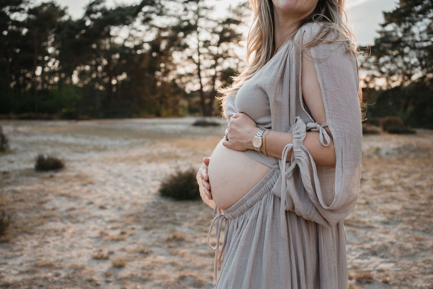 Detailopname van een zwangere blote buik, liefdevol vastgehouden door een vrouw in grijze jurk op een zandvlakte in de natuur van Soest.