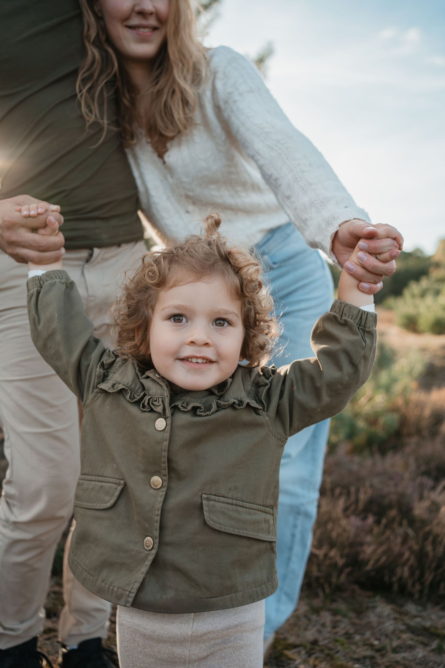 Ouders houden de handjes van hun dochter vast tijdens een gezinsfotoshoot in de natuur.