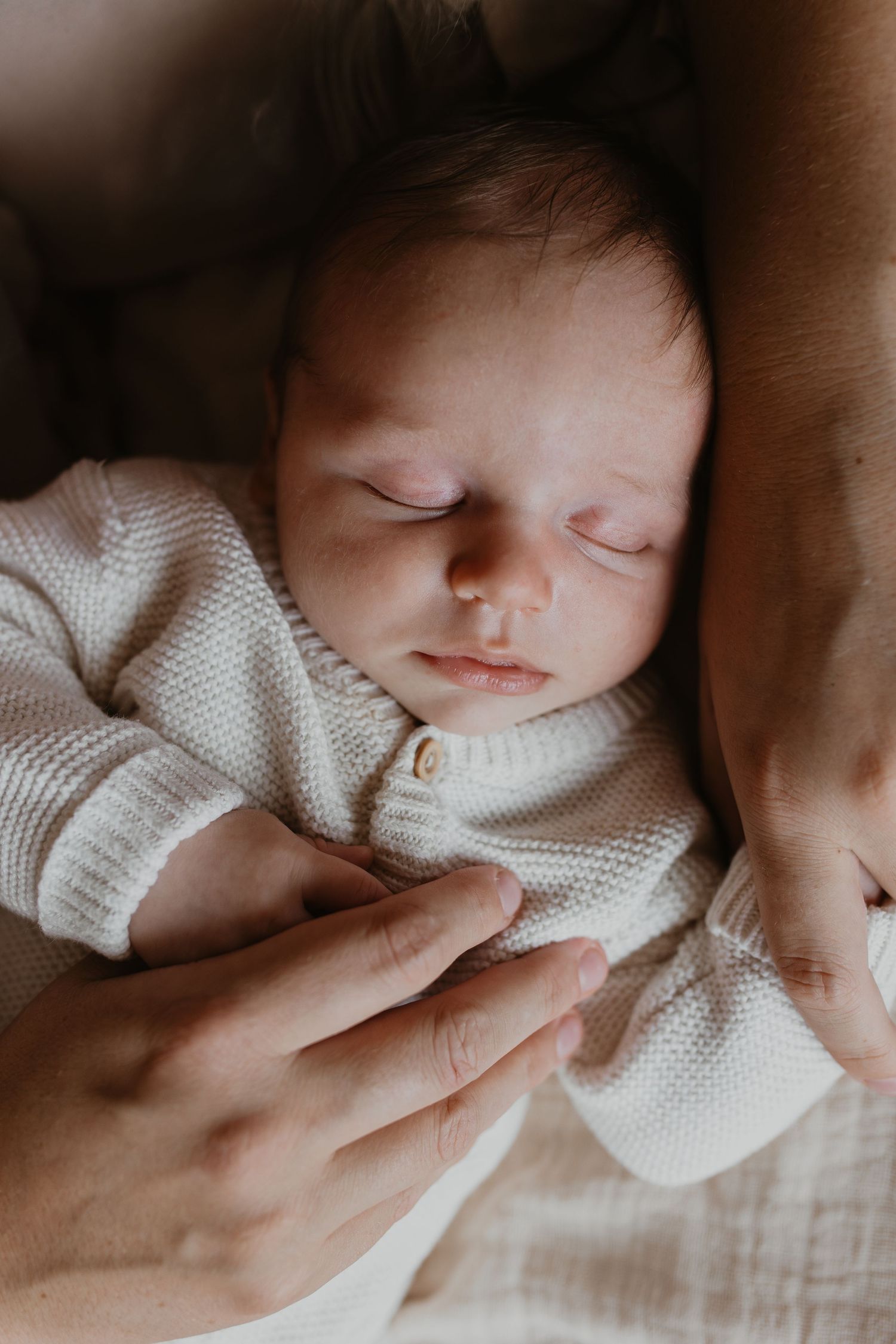 Close up van slapende newborn baby in een gebreid box pakje, omringd door de handen van een ouder tijdens de newbornshoot.
