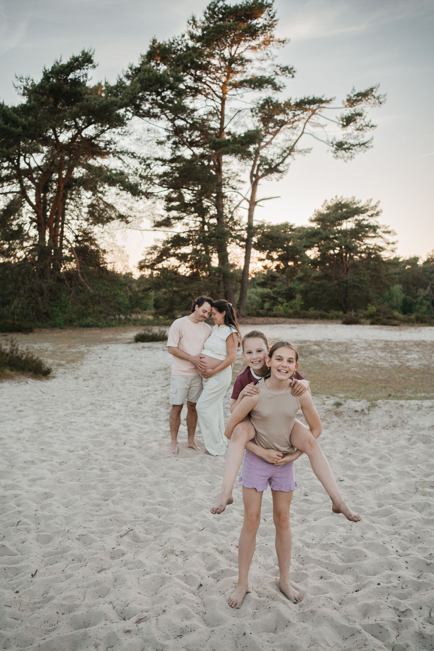 Een lachend meisje geeft haar zusje een piggyback-ritje, met hun zwangere ouders liefdevol op de achtergrond, in de Soester Duinen.