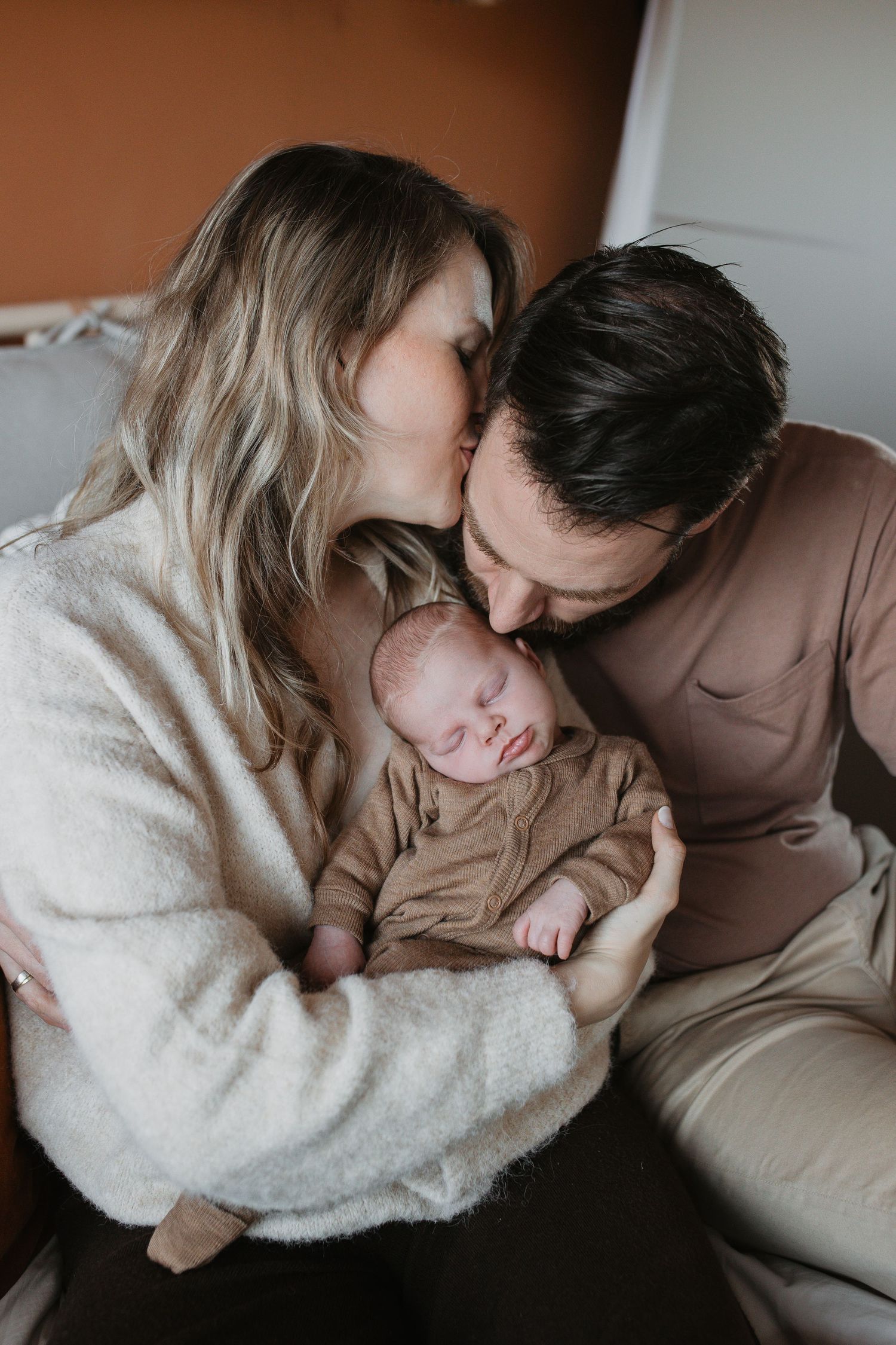 Prachtige newbornshoot in warme aardetinten. Mama kust papa en papa kust de baby, die comfortabel en slapend tussen hen in ligt.