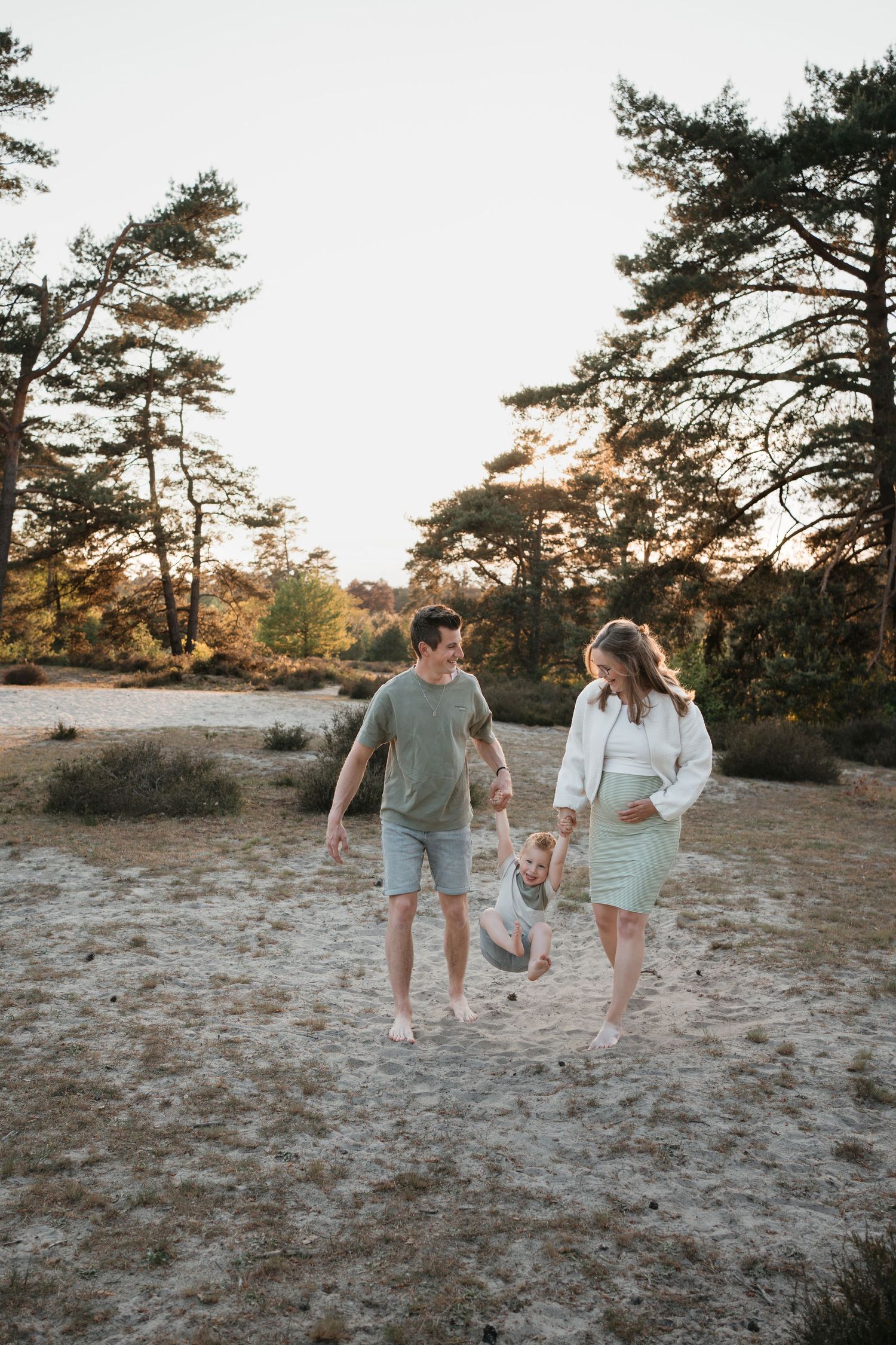 Zwangere vrouw en haar man in groene kleding wandelen hand in hand met hun lachende kind tijdens een fotoshoot in de natuur van Soest.
