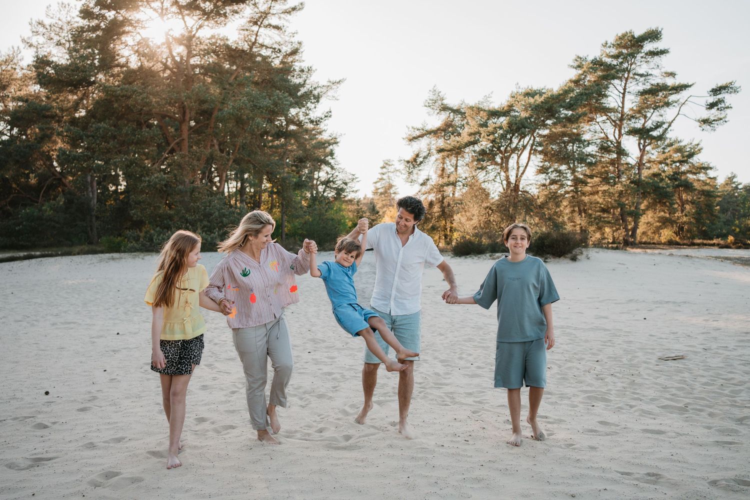 Een kleurrijk gezin wandelt hand in hand door het zand van de Soester Duinen, terwijl de ouders hun jongste zoon speels zwieren.