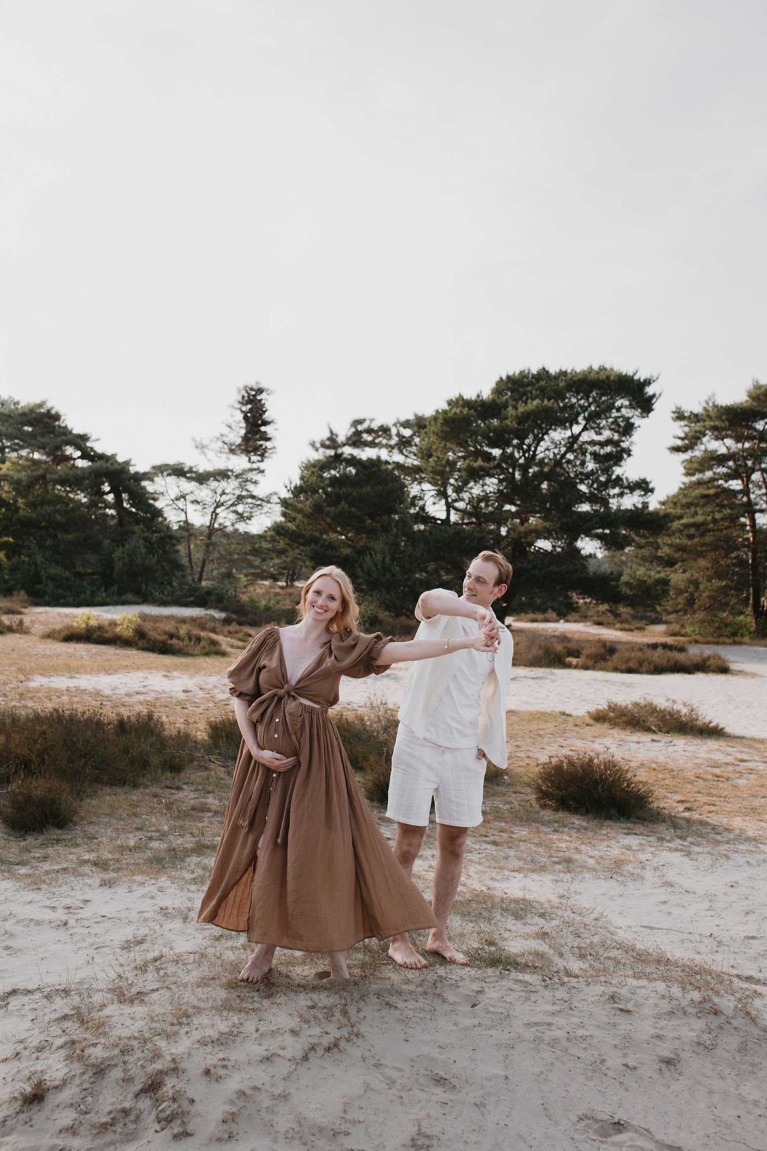 Man en vrouw staat in bosrijk gebied in het zand. Bewegen alsof zij aan het dansen zijn. De vrouw maakt een draai en kijkt naar de camera.