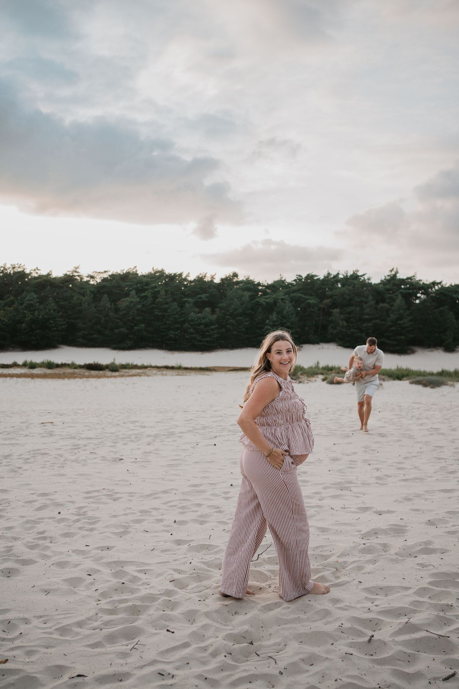 Zwangere vrouw glimlacht in het zand, op de achtergrond speelt een man met een kindje.