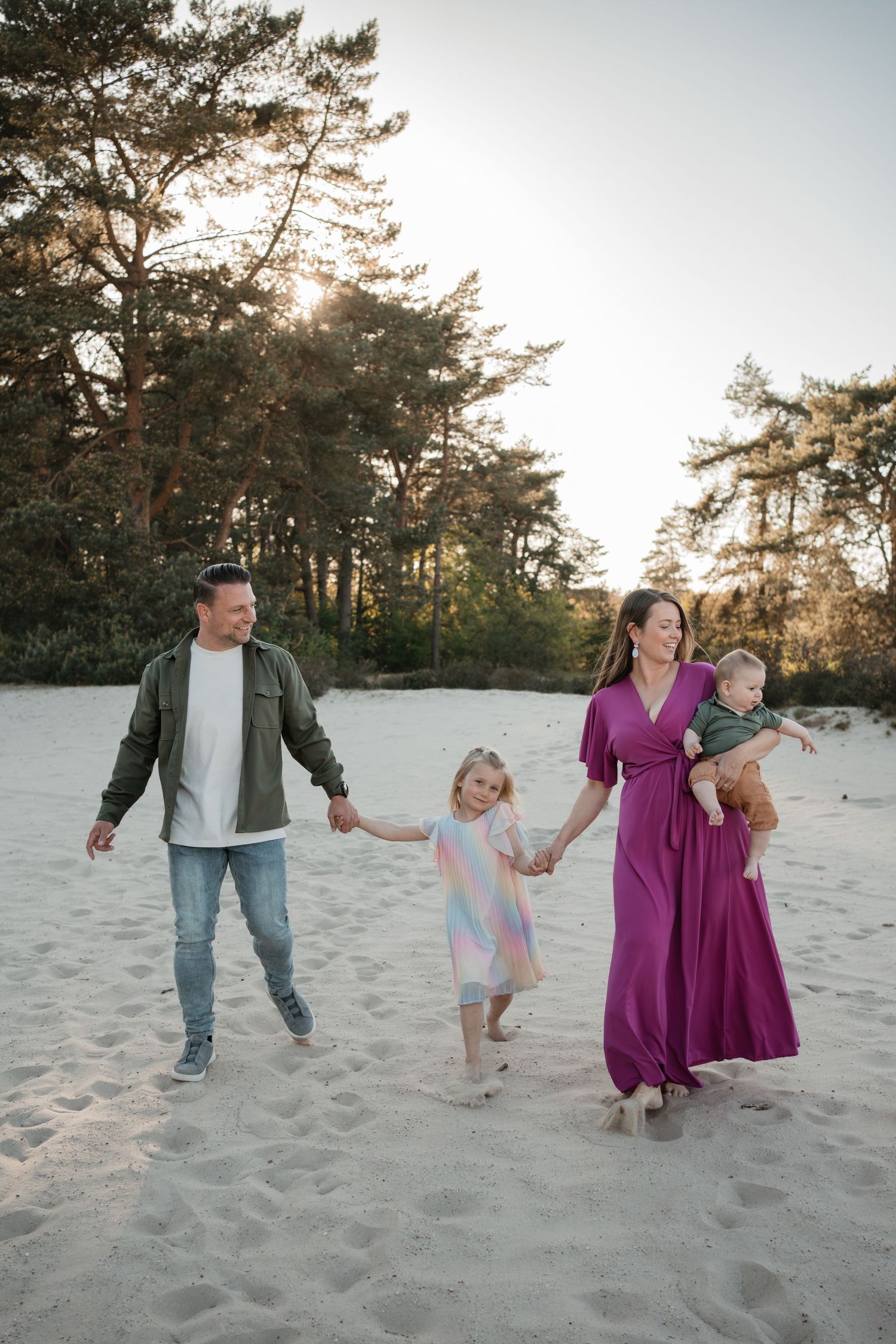 Spontane gezinsshoot in de natuur bij zonsondergang met een vrolijke vader, moeder in paarse jurk, dochter en baby in het zand.