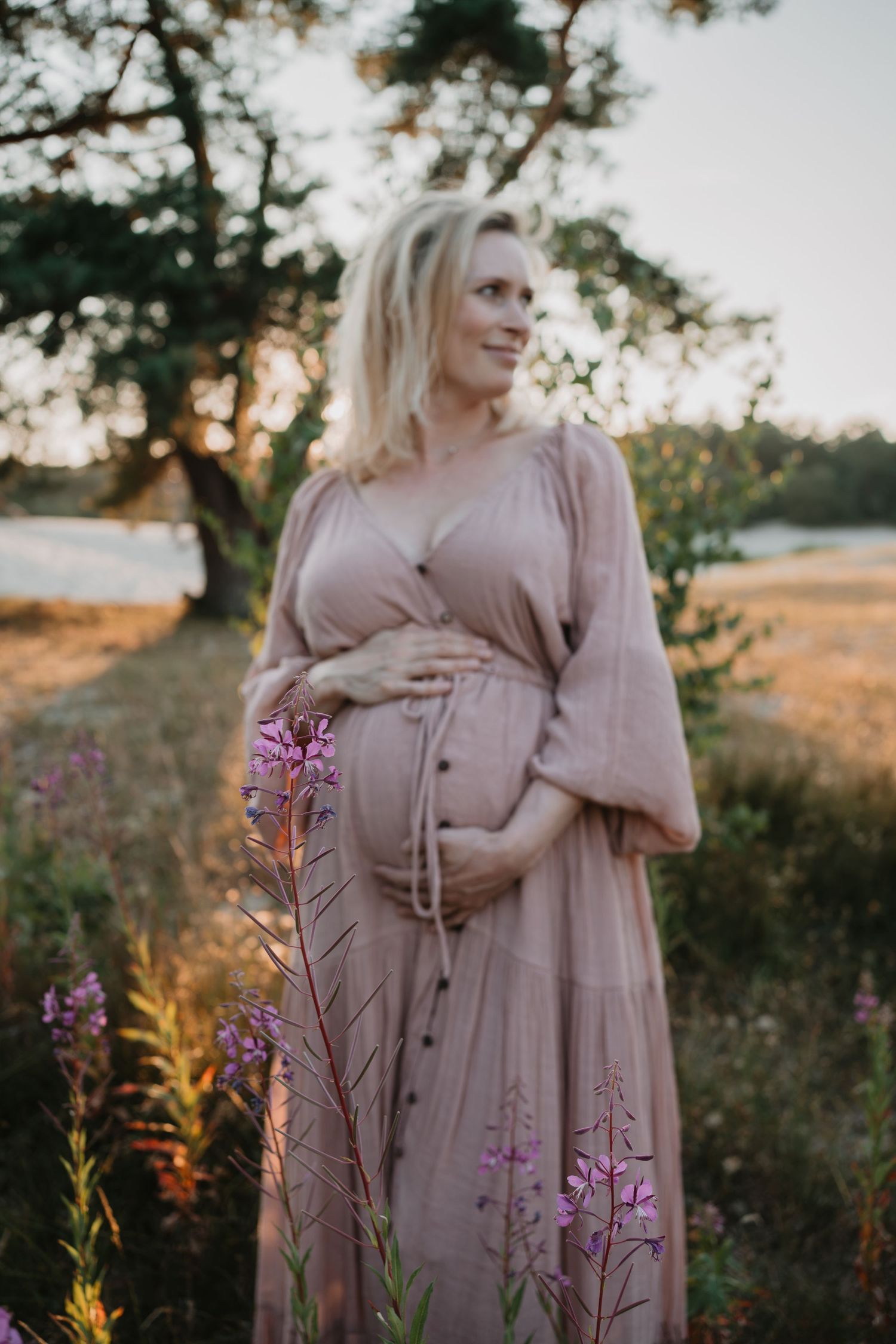 Zwangere vrouw in lichtroze jurk poseert tijdens een gouden zonsondergang tussen wilde bloemen in een natuurlijke omgeving.