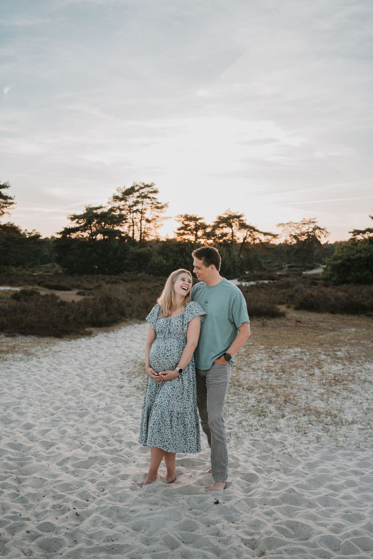 Een zwanger stel lacht samen op een zandvlakte in de natuur van Soest,Soester Duinen tijdens een fotoshoot bij een sfeervolle zonsondergang.