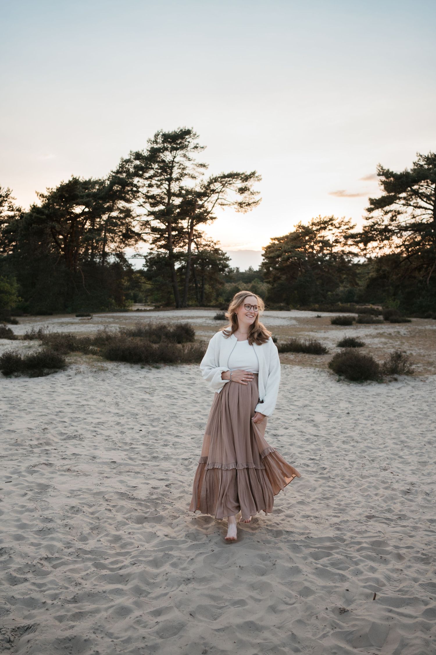 Een zwangere vrouw met blond haar loopt glimlachend op blote voeten op de Soester Duinen. Ze draagt een lange rok en een wit vest.