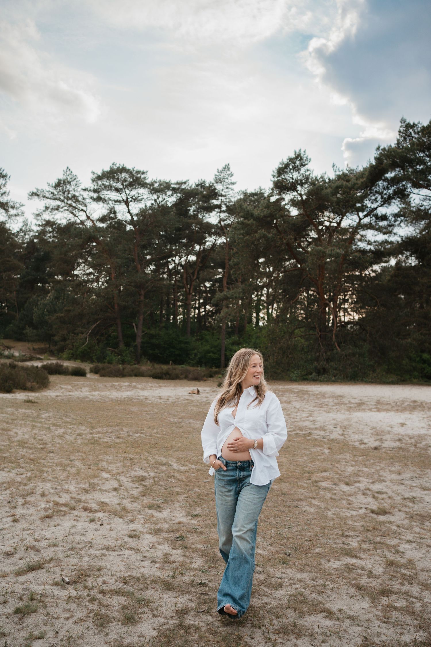 Zwangerschapsshoot op de Soester Duinen. Zwangere vrouw in witte open blouse met spijkerbroek houdt liefdevol haar blote buik vast.