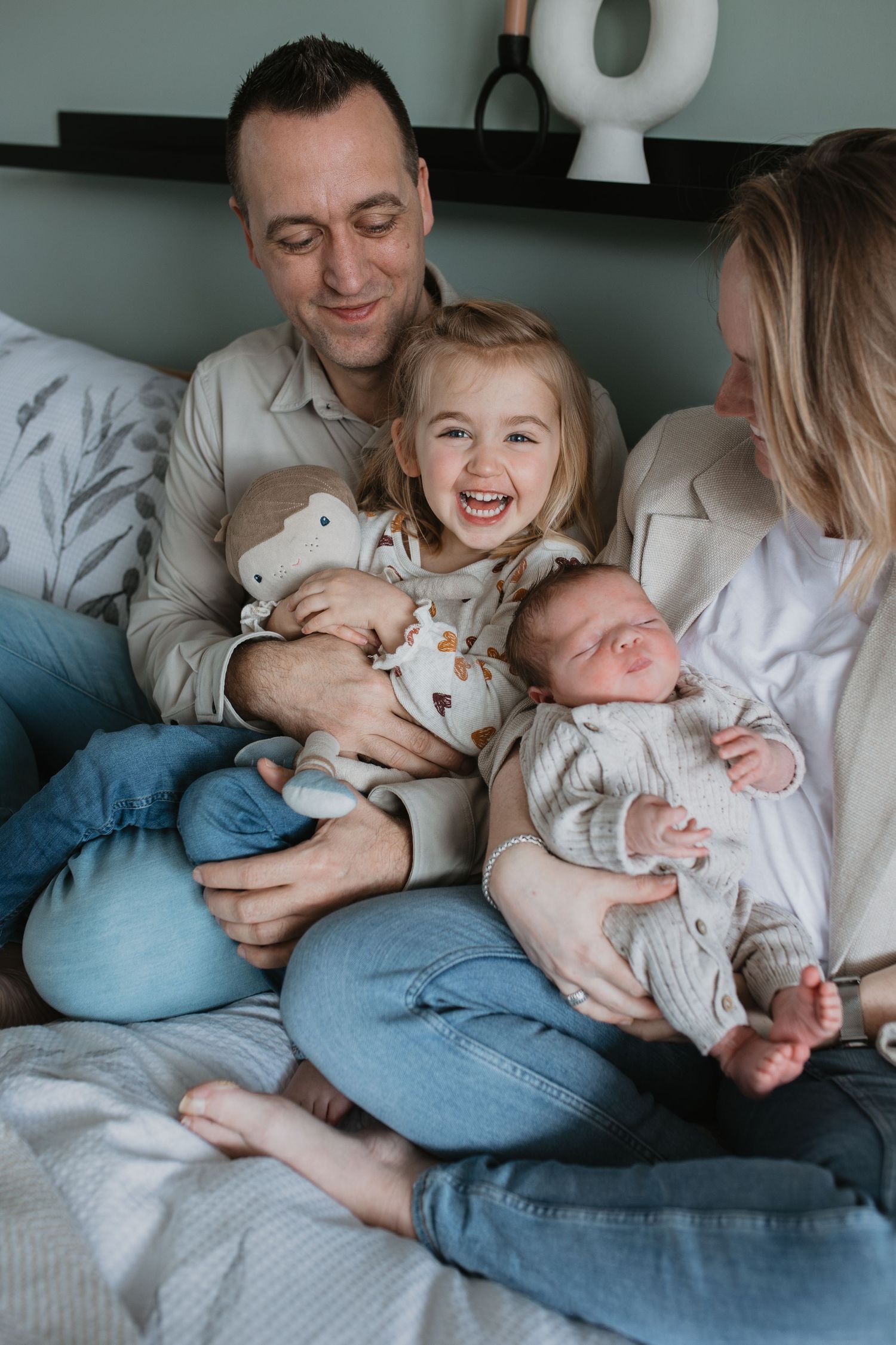 Ouders knuffelen op bed met hun jonge, vrolijke dochter kijkt in de lens en een pasgeboren baby tijdens een gezinsfotoshoot