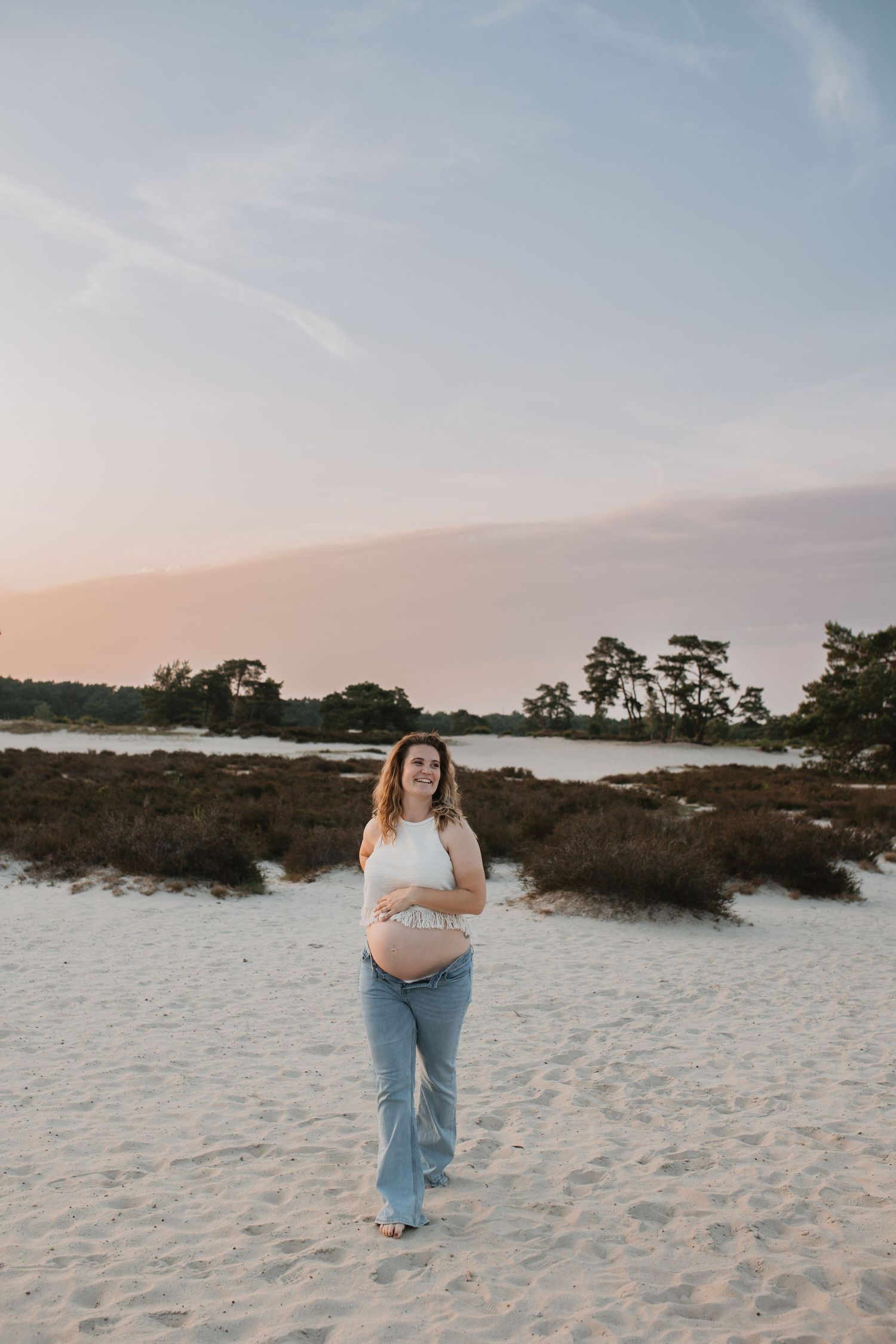 Zwangere vrouw loopt lachend richting de voorgrond. Haar buik is ontbloot, zij is duidelijk zwanger.