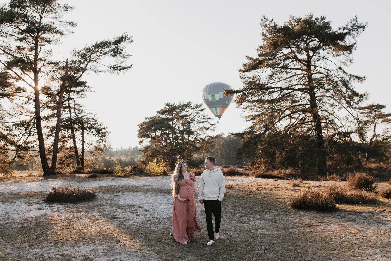Zwanger stel loopt hand in hand over een duin- en bosrijk gebied. Op de achtergrond zweeft een luchtballon.