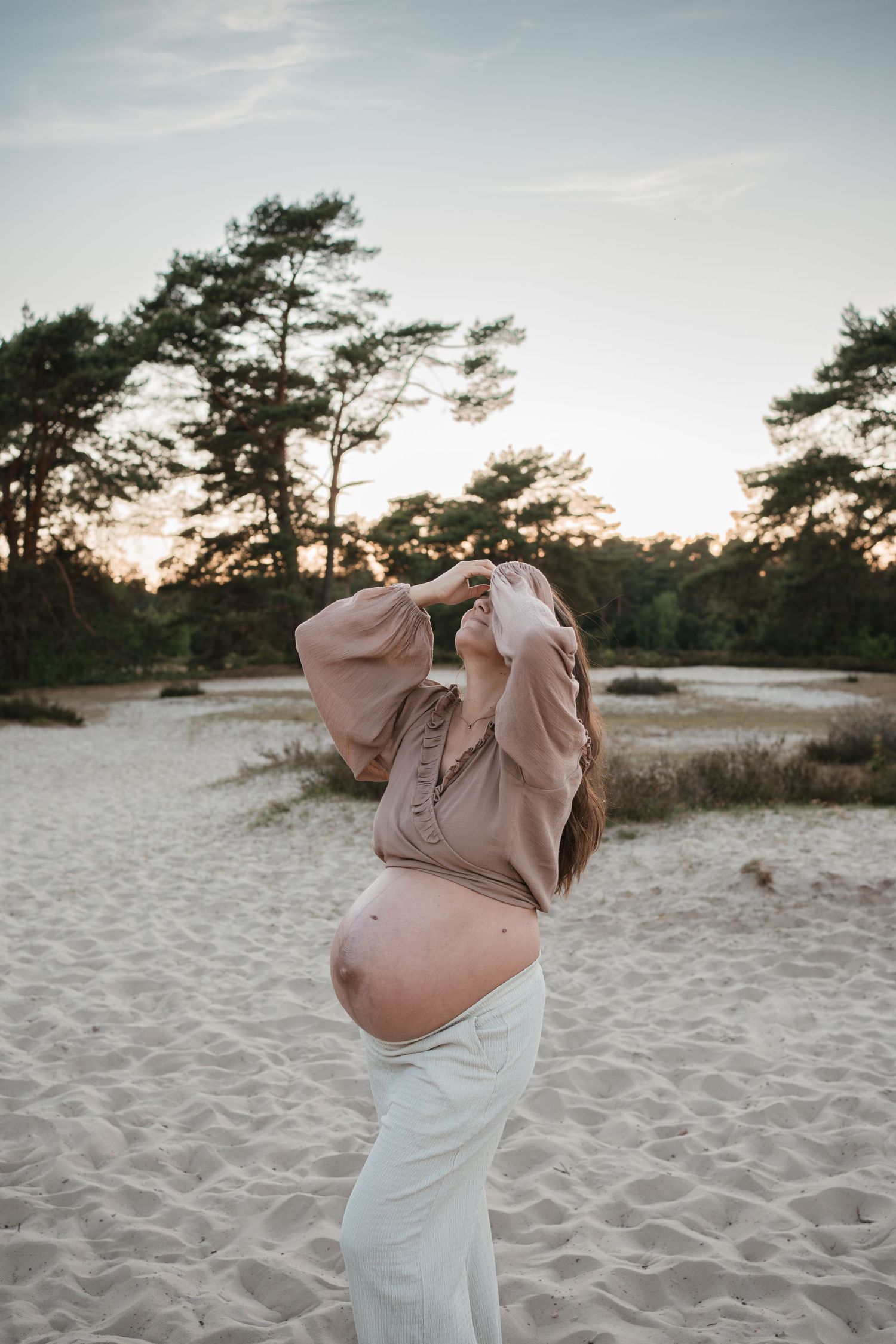Zwangere vrouw met blote buik staat met haar armen omhoog en handen voor haar gezicht in de Soester Duinen.
