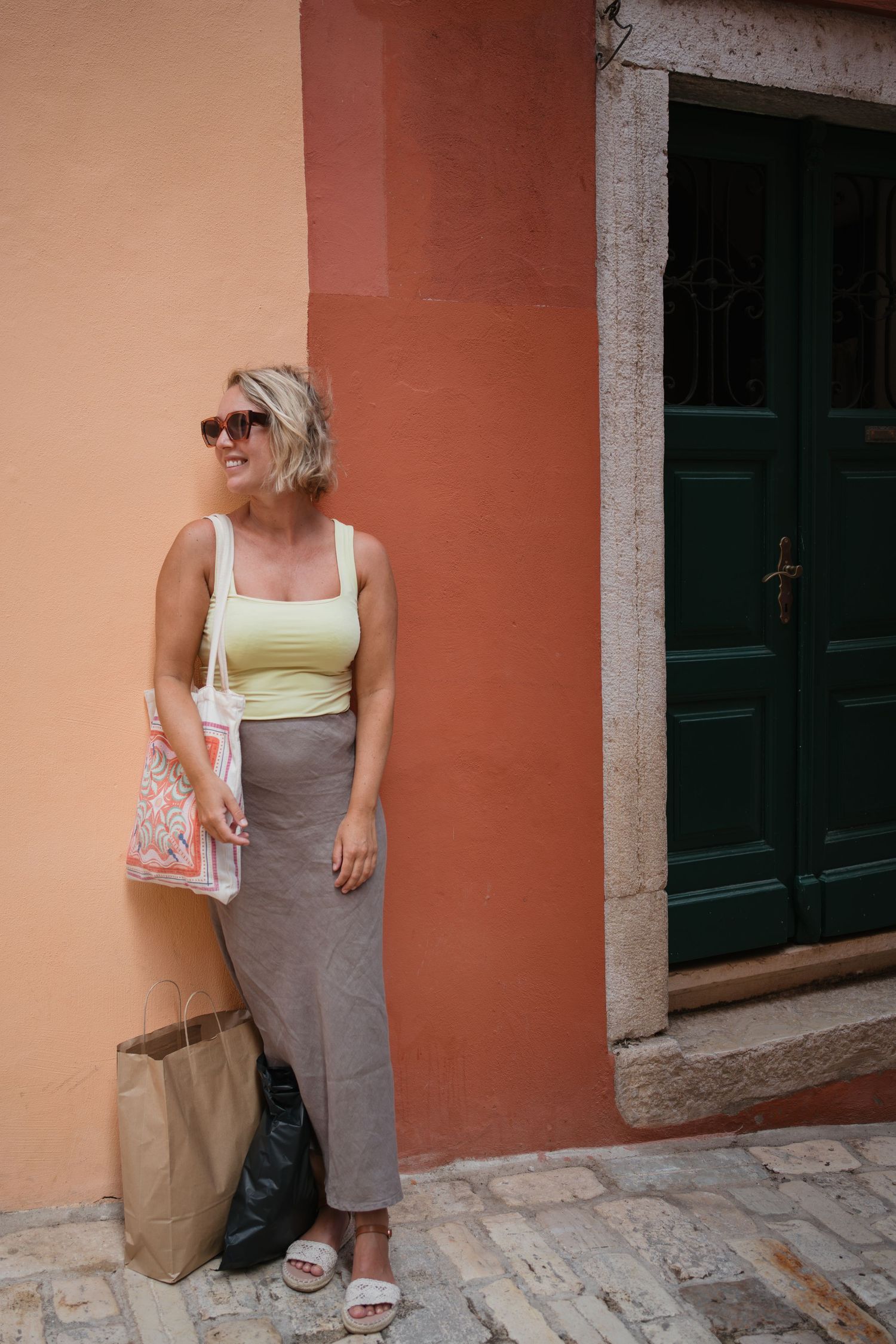 Lachende blonde vrouw met zonnebril en tas leunt tegen een warme oranje muur naast een donkergroene deur.