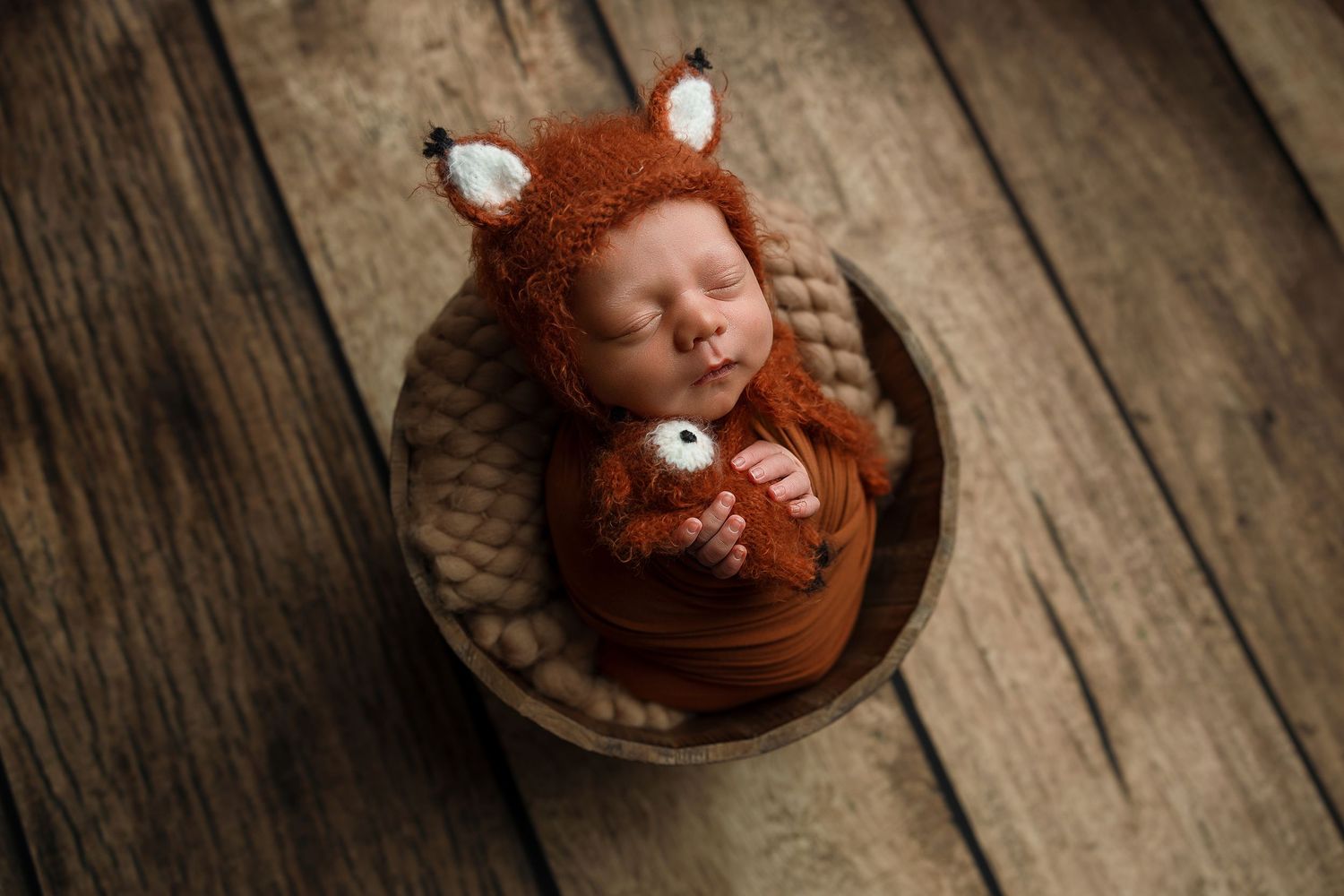 Newborn baby wrapped in orange knit swaddle wearing fox bonnet, sleeping in wooden bowl on rustic wood floor.
