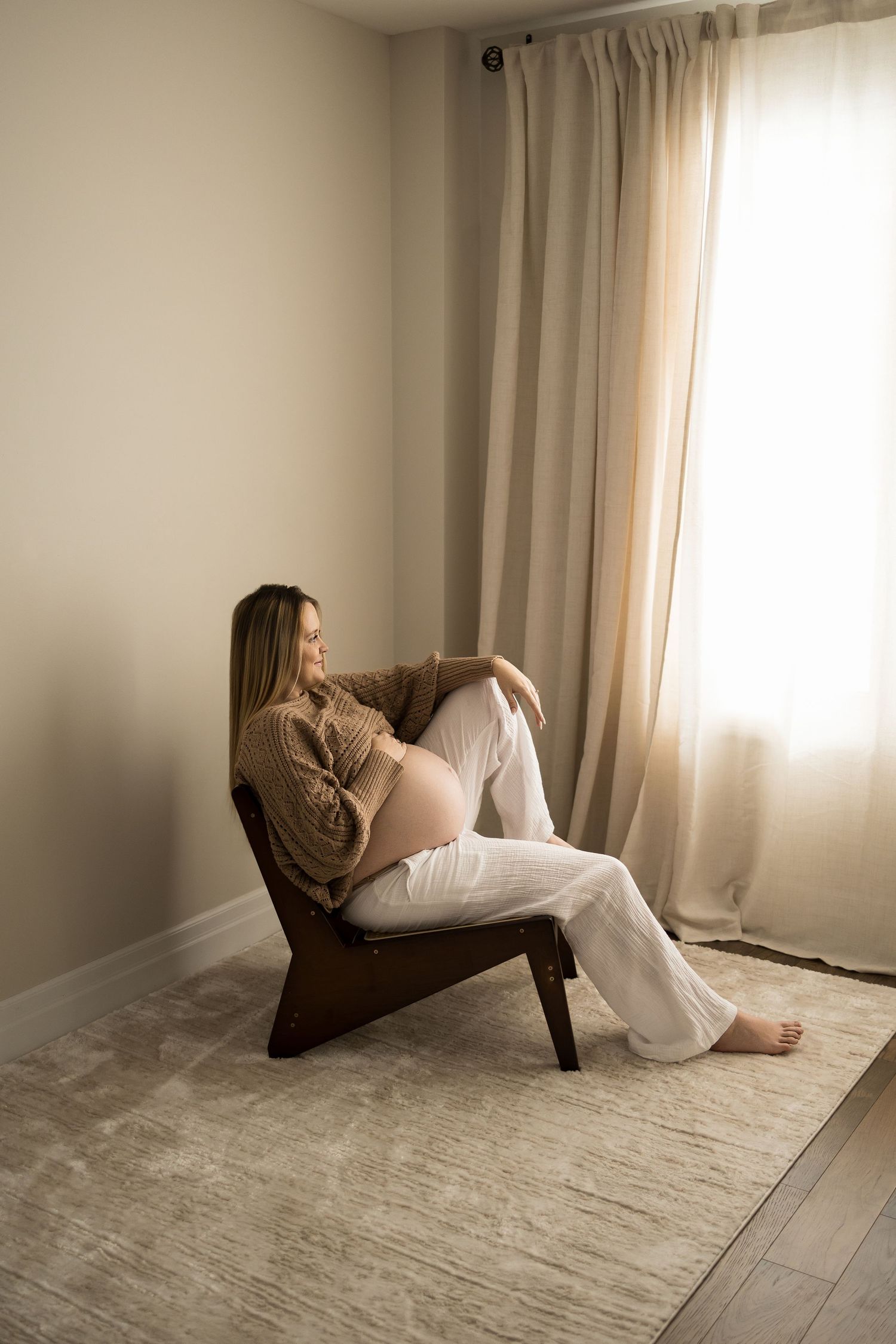 A pregnant woman relaxes in a modern wooden chair by a window with sheer curtains in a minimalist room.
