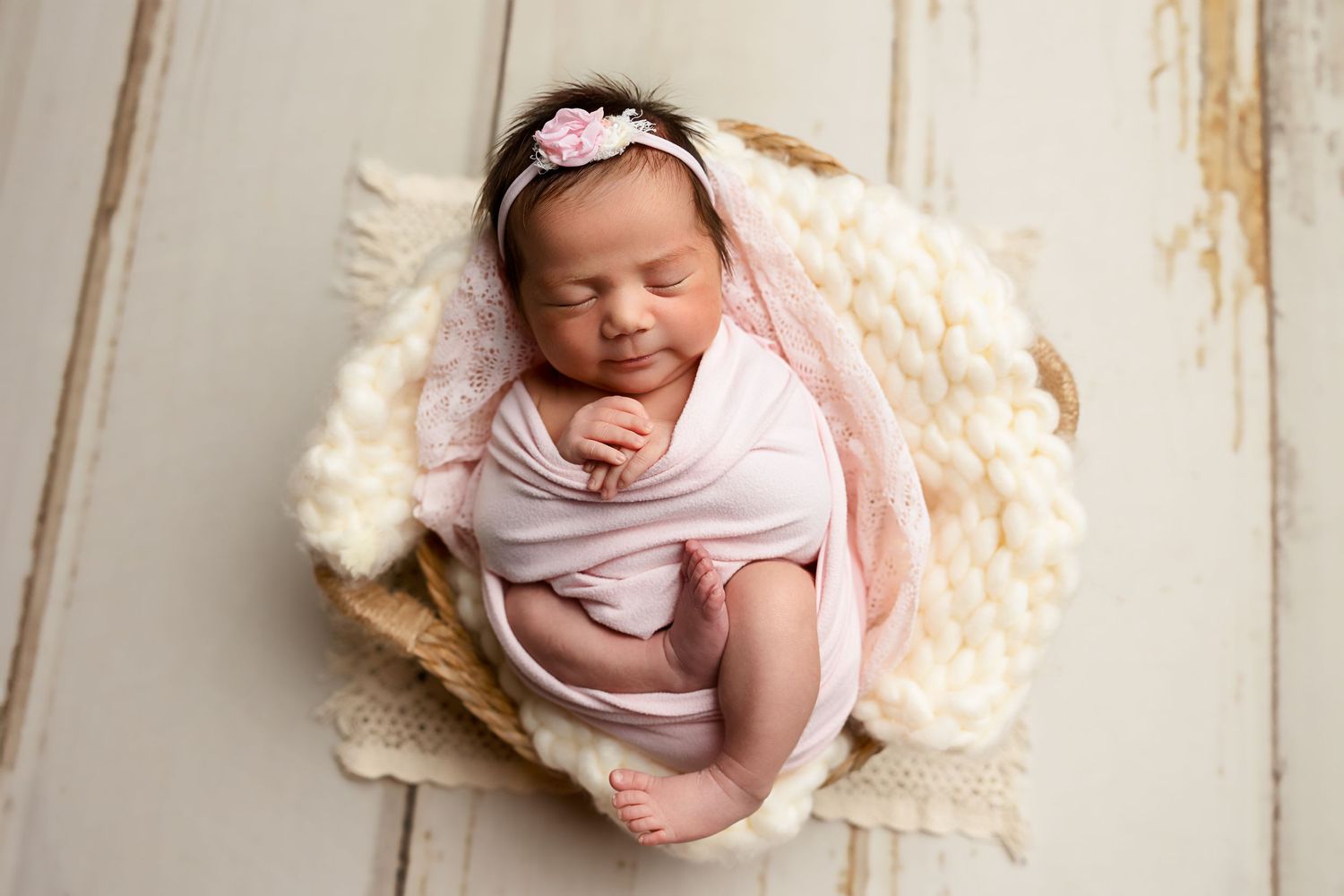 Sleeping newborn wrapped in pink swaddle resting in cozy cream basket on rustic wooden background.
