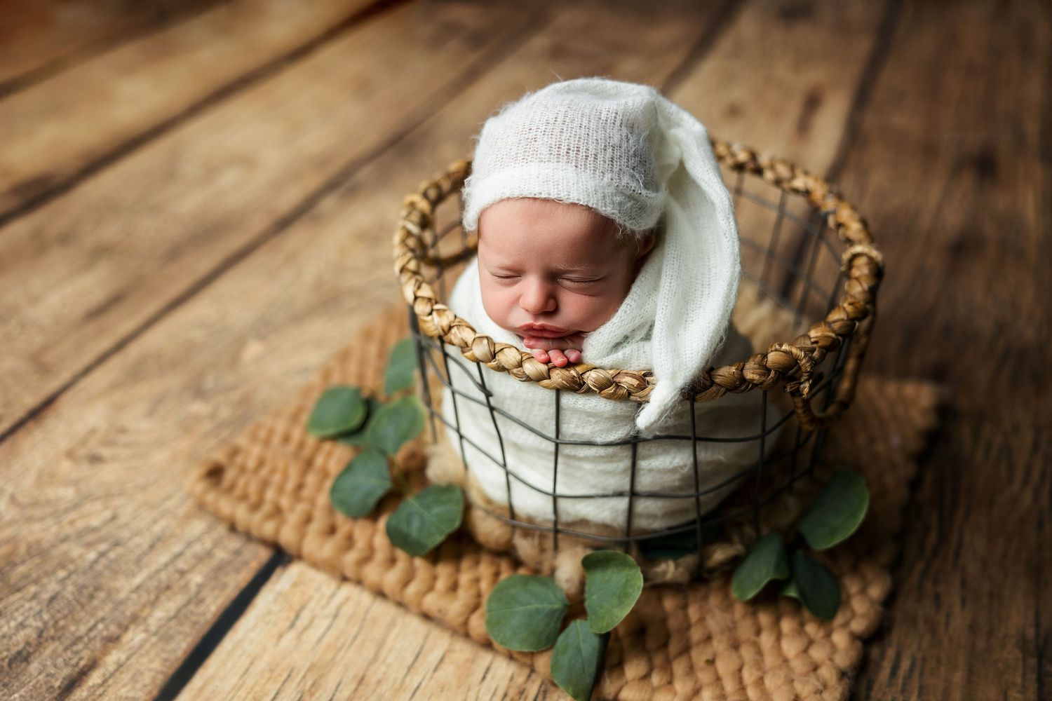 Sleeping newborn in white hat nestled in wire basket with greenery on wooden floor.