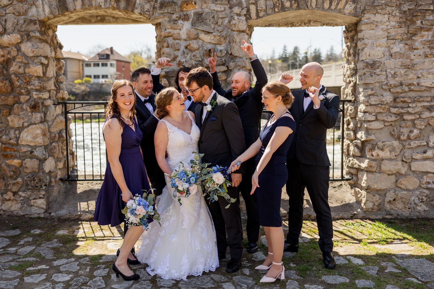 Wedding party poses playfully between stone archways during outdoor celebration.