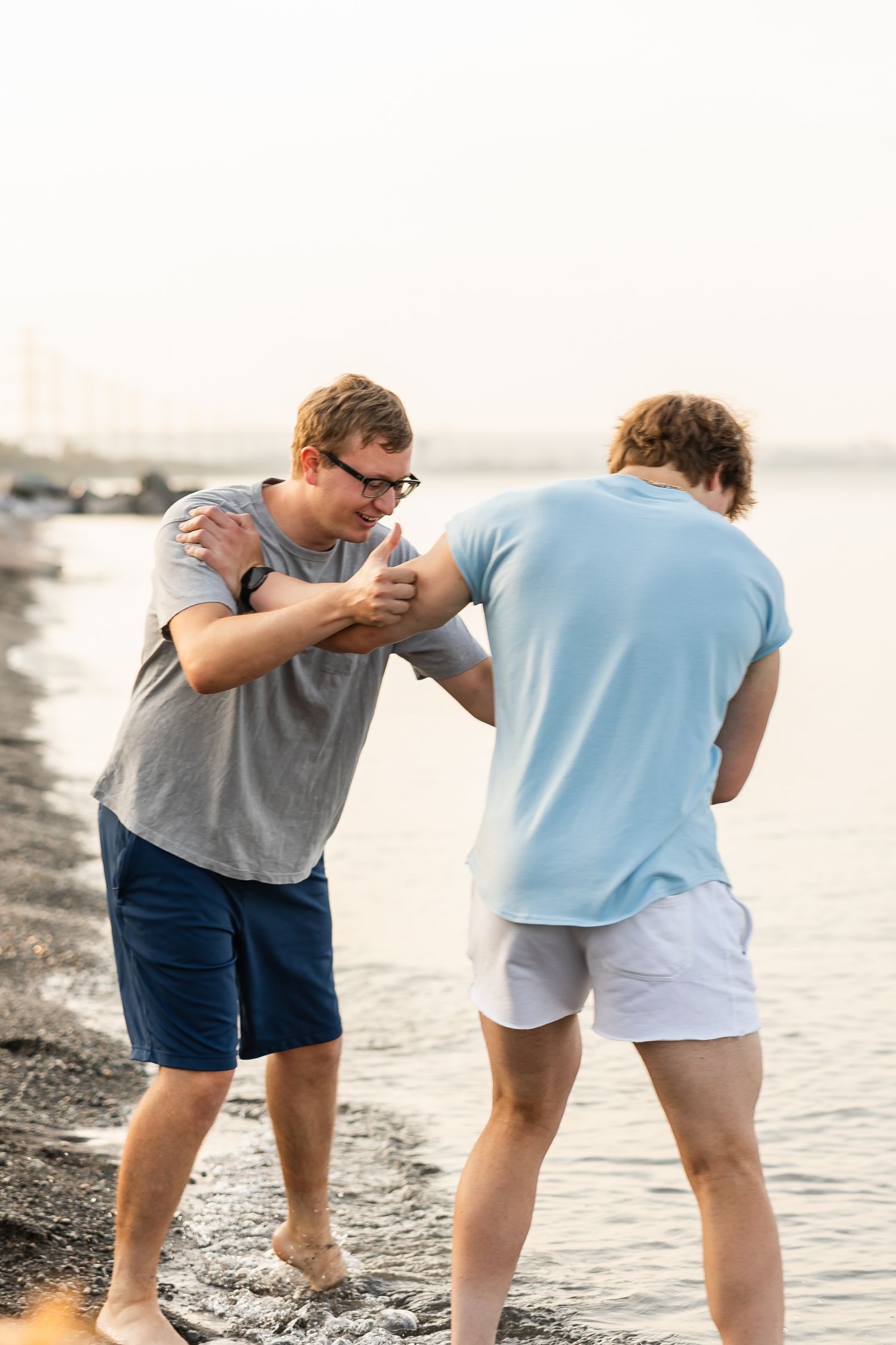 Two men playfully push each other while standing barefoot on a beach at sunset.