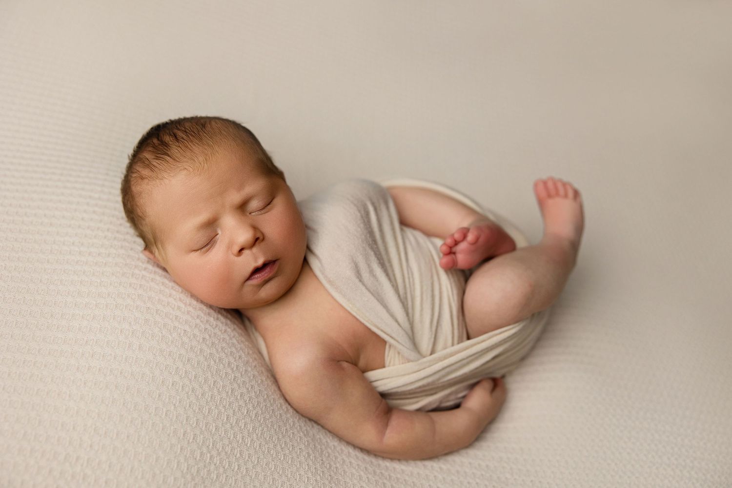 Sleeping newborn wrapped in soft white fabric lying on textured cream backdrop during studio photo session.