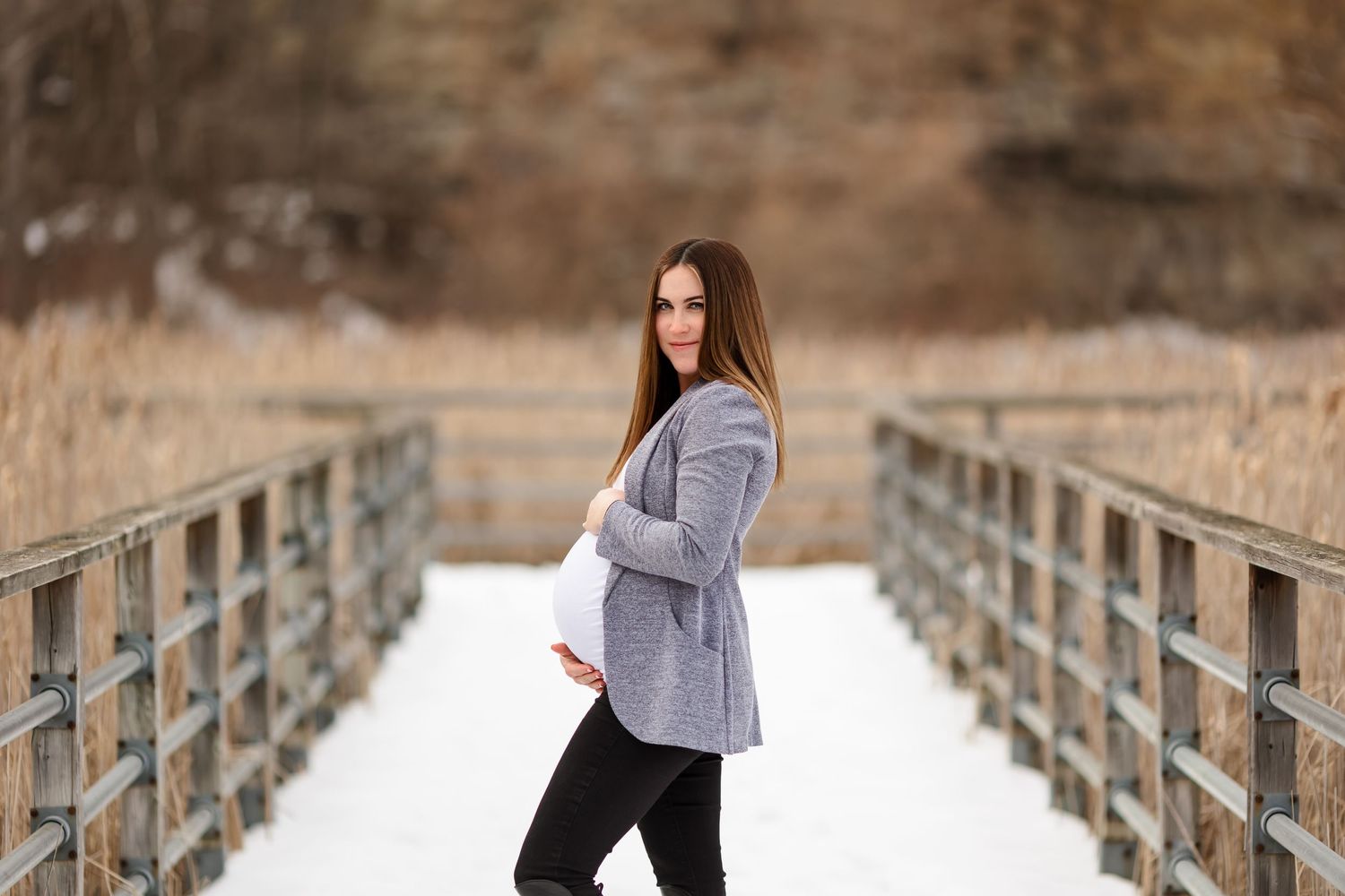 Woman in grey cardigan poses on snowy bridge during winter maternity photoshoot.