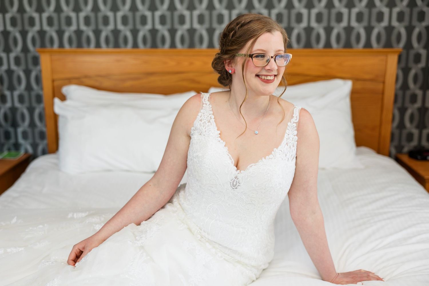 A person in a white wedding dress sits elegantly on a bed with a patterned headboard.