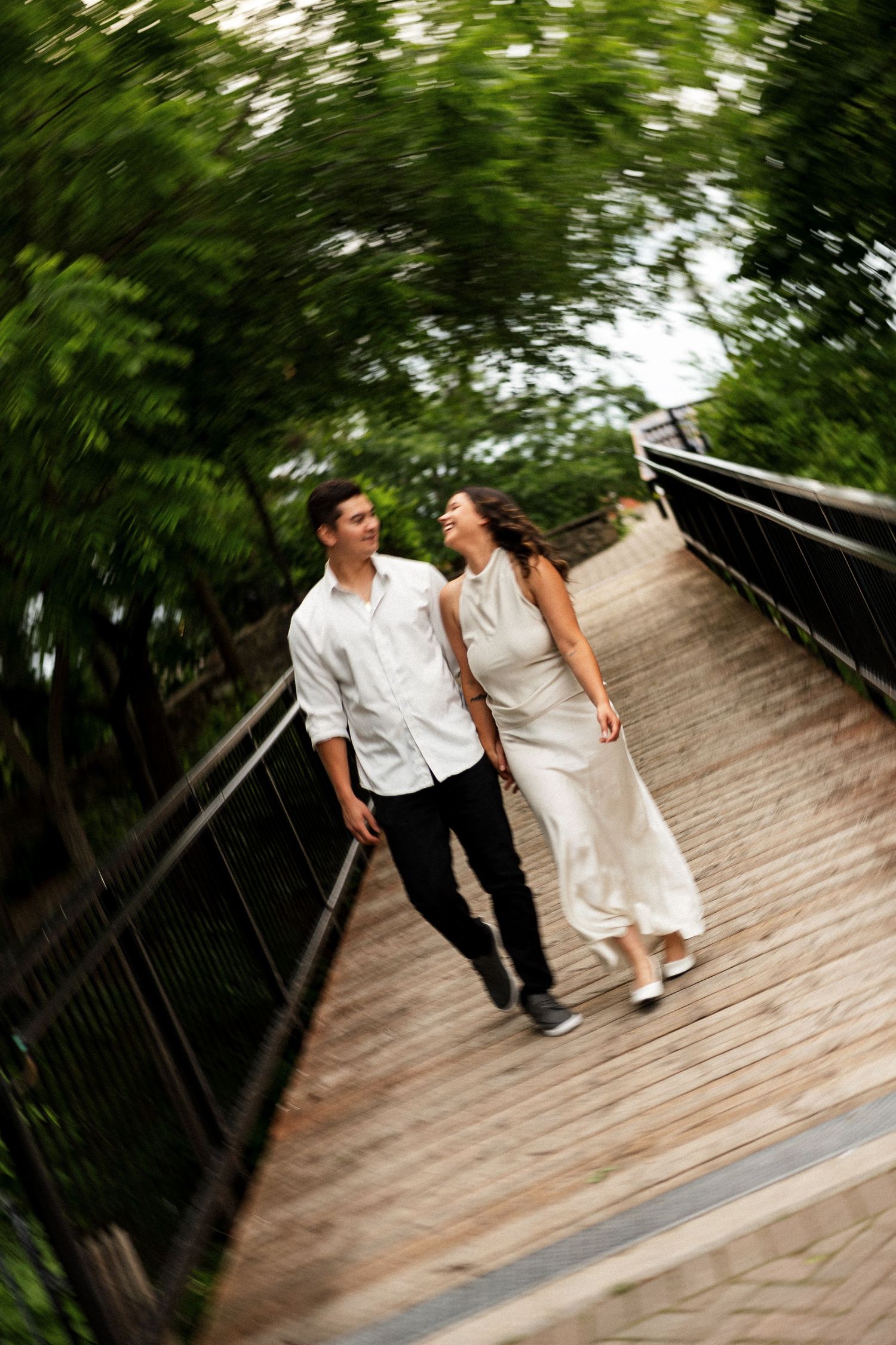 A couple walks hand-in-hand on a wooden bridge surrounded by lush green trees, smiling at each other.