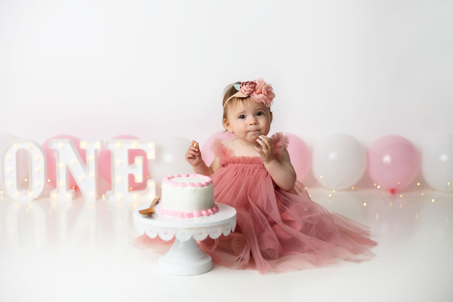 Pink themed first birthday setup with tulle dress, illuminated ONE sign and white balloons.