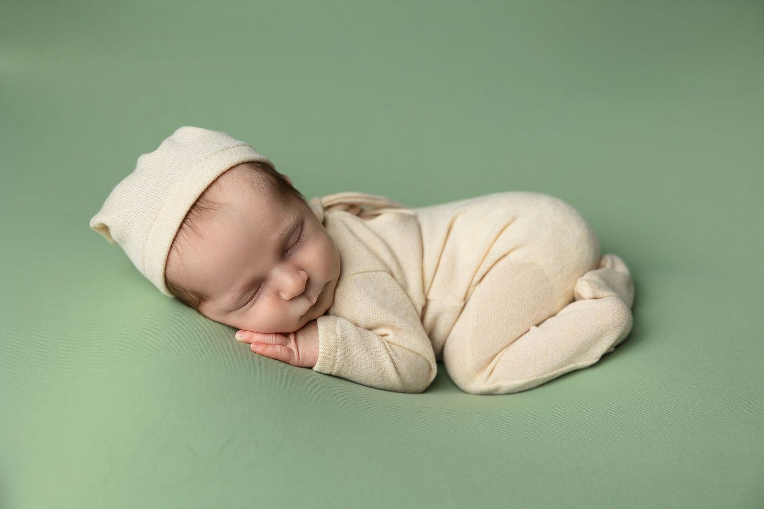 Sleeping newborn in cream outfit lying peacefully on sage green backdrop.