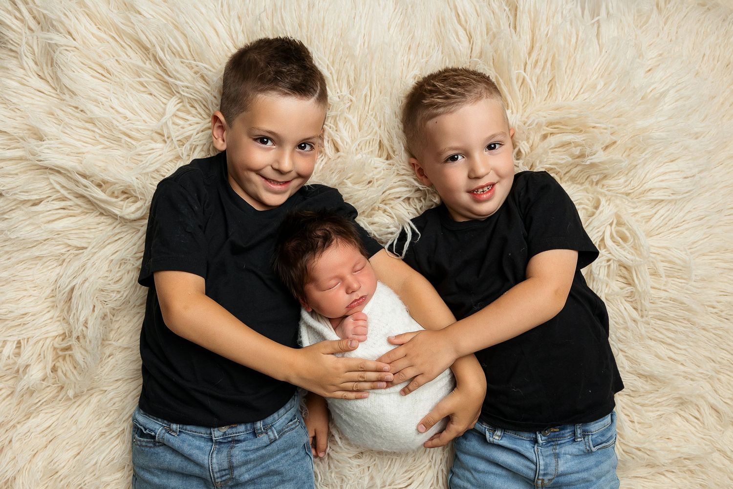 Siblings in matching black shirts pose playfully together on a cream-colored fur rug for a family photo session.
