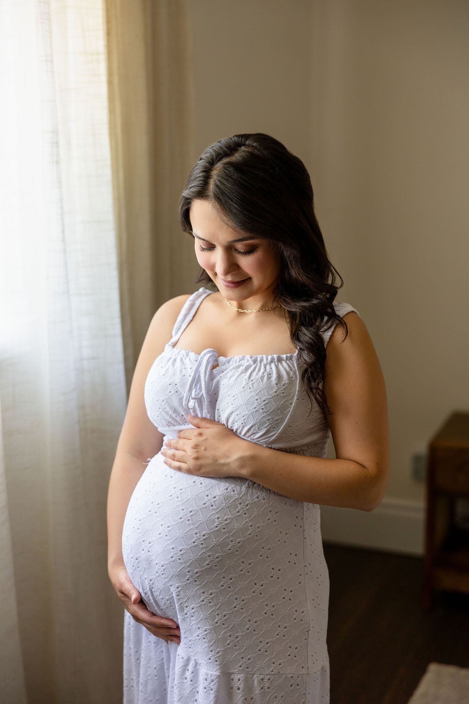 A person in a white eyelet lace dress poses near a window during a maternity photoshoot.