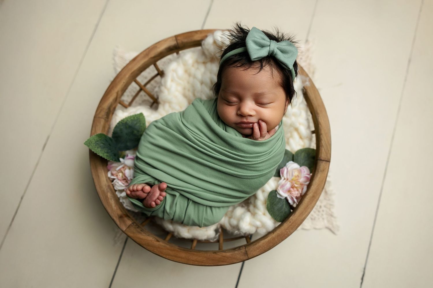 Sleeping newborn wrapped in mint green swaddle rests in wooden bowl with floral accents.