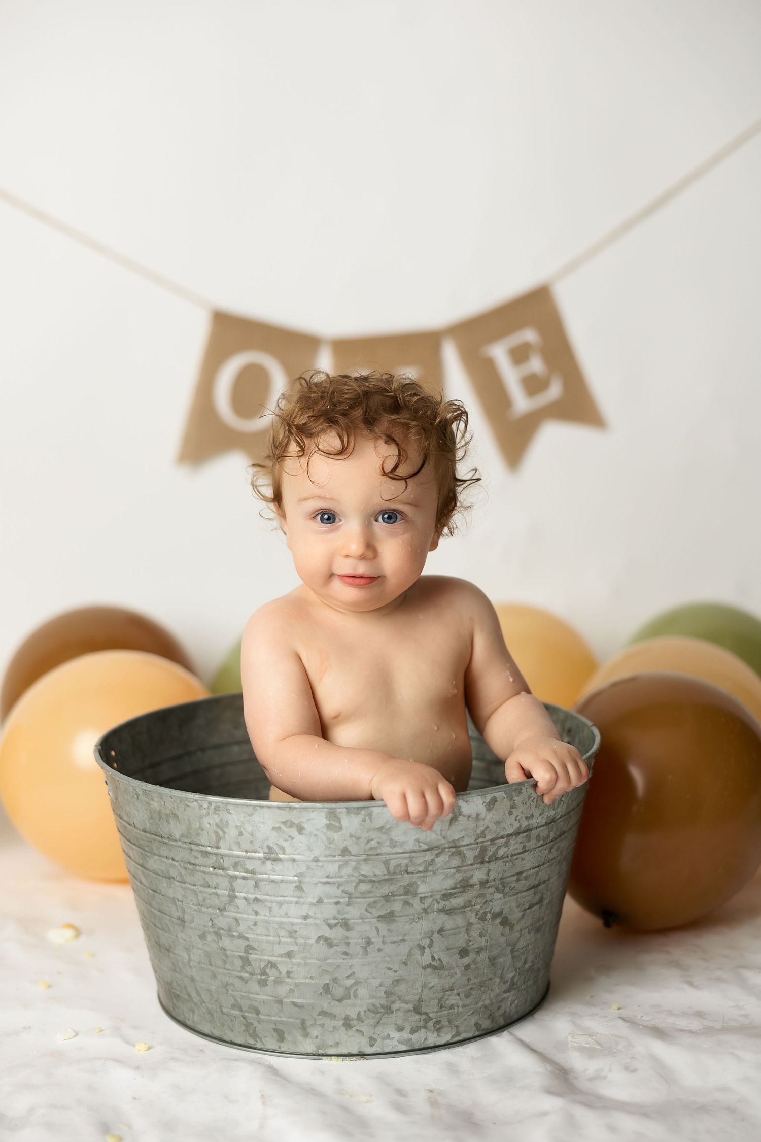 Smiling toddler in galvanized tub with ONE banner and earth-toned balloons behind.
