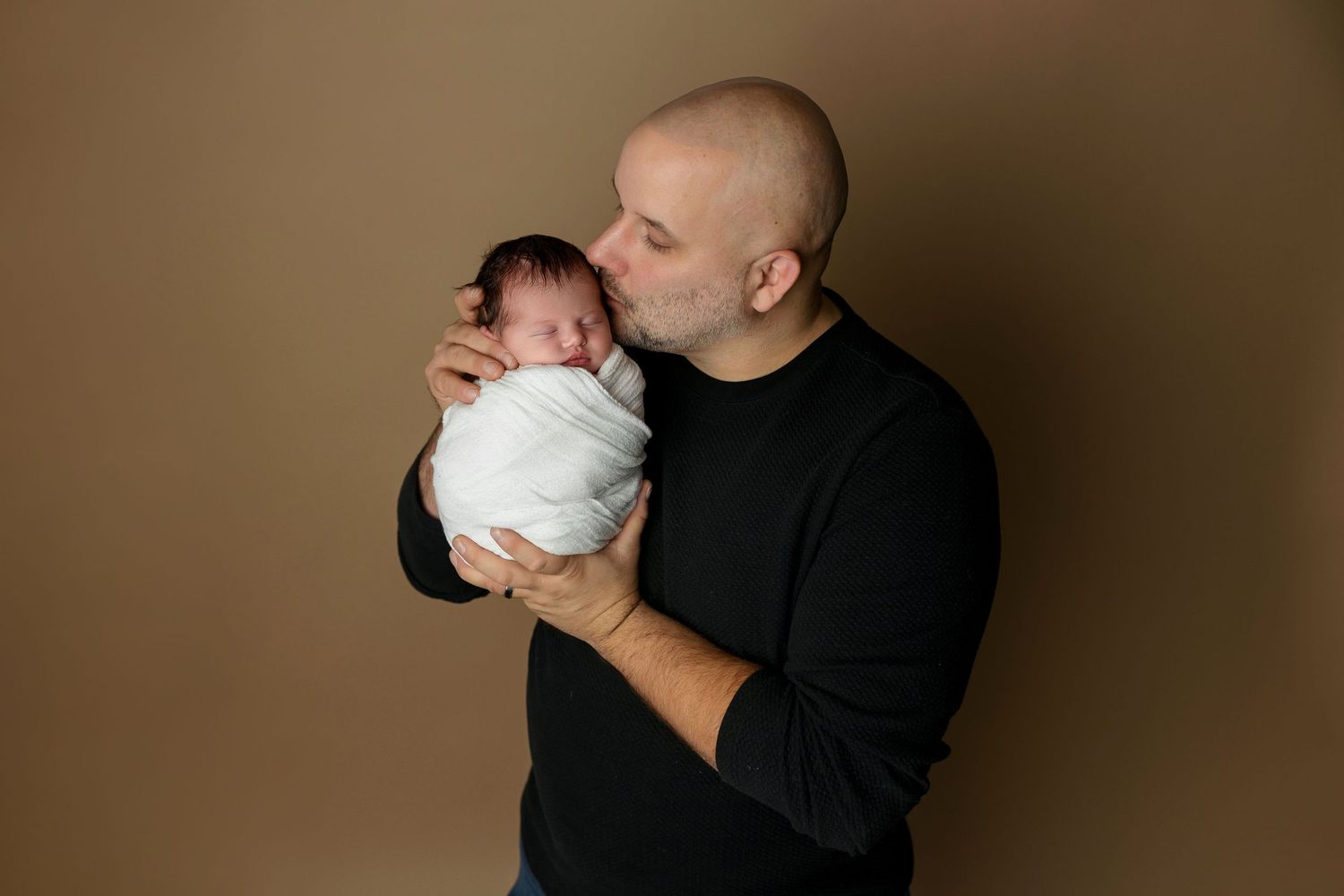 Tender moment between parent and swaddled newborn against warm brown studio background.