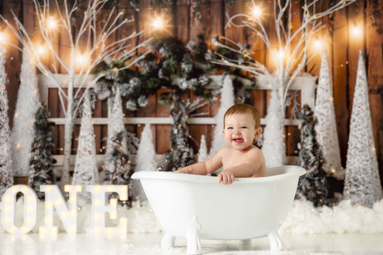 Smiling baby in white bathtub with winter wonderland backdrop featuring lit trees and ONE sign.