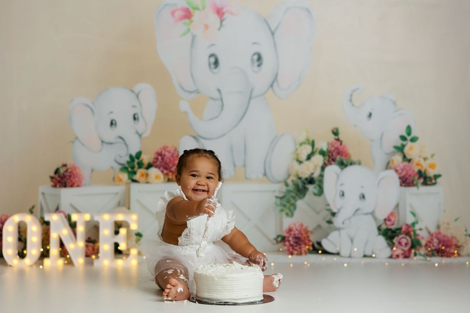 First birthday setup with illuminated ONE sign and whimsical elephant backdrop with floral decorations.