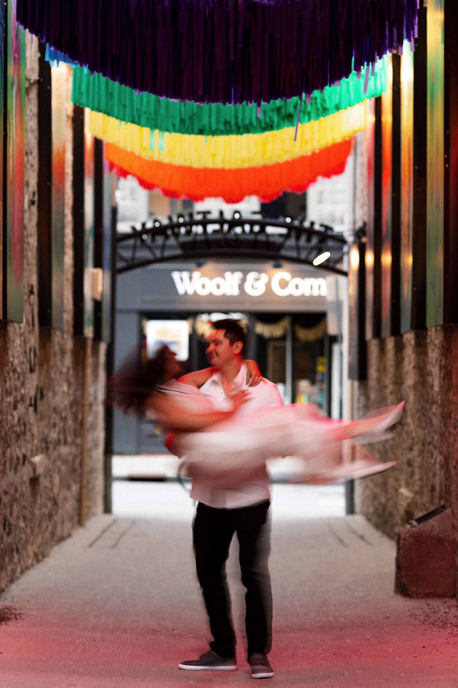 Couple dancing under rainbow fringe decorations outside Woolf & Com store at night.
