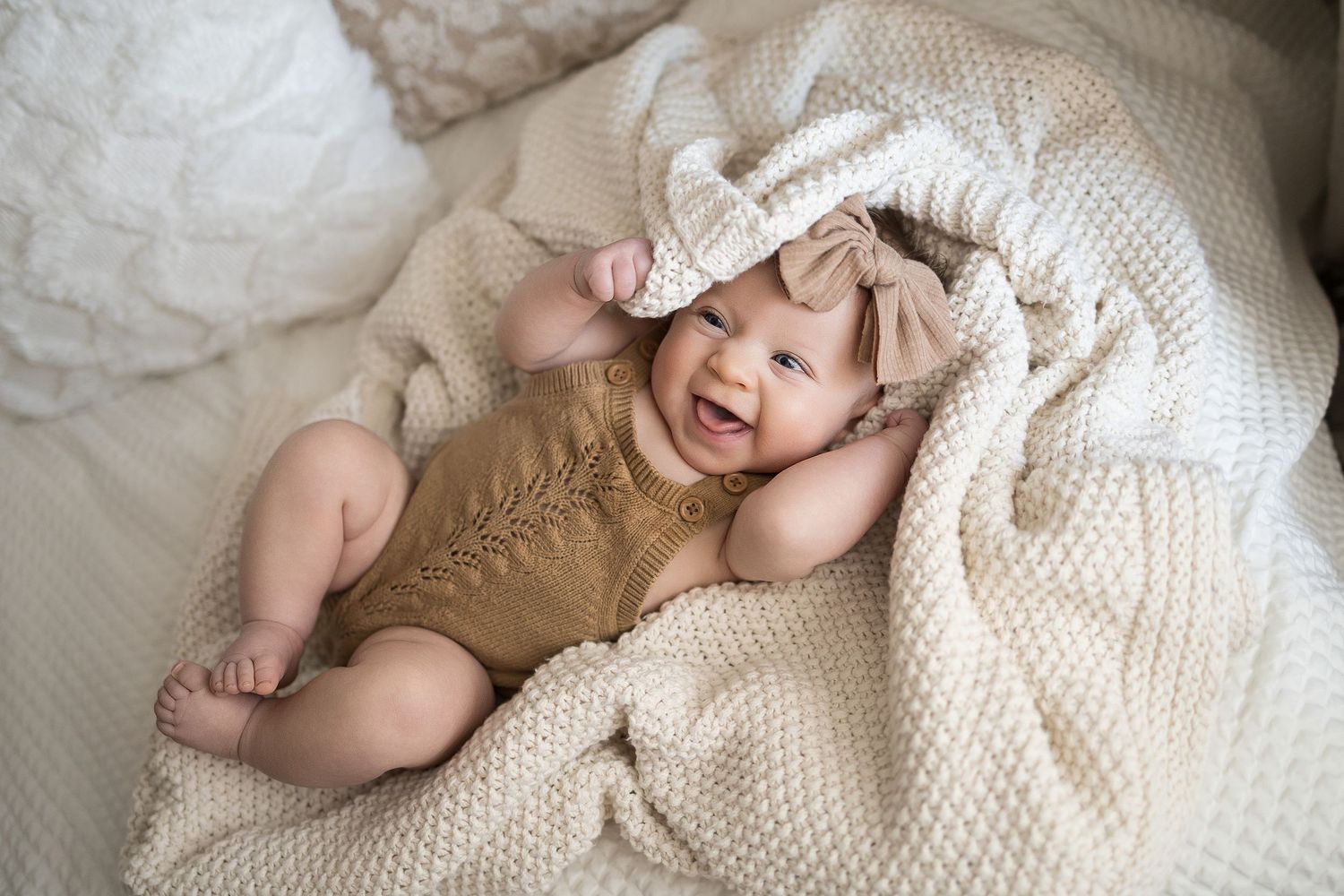 Smiling baby in brown knit romper and white bow headband lying on cream knit blanket.