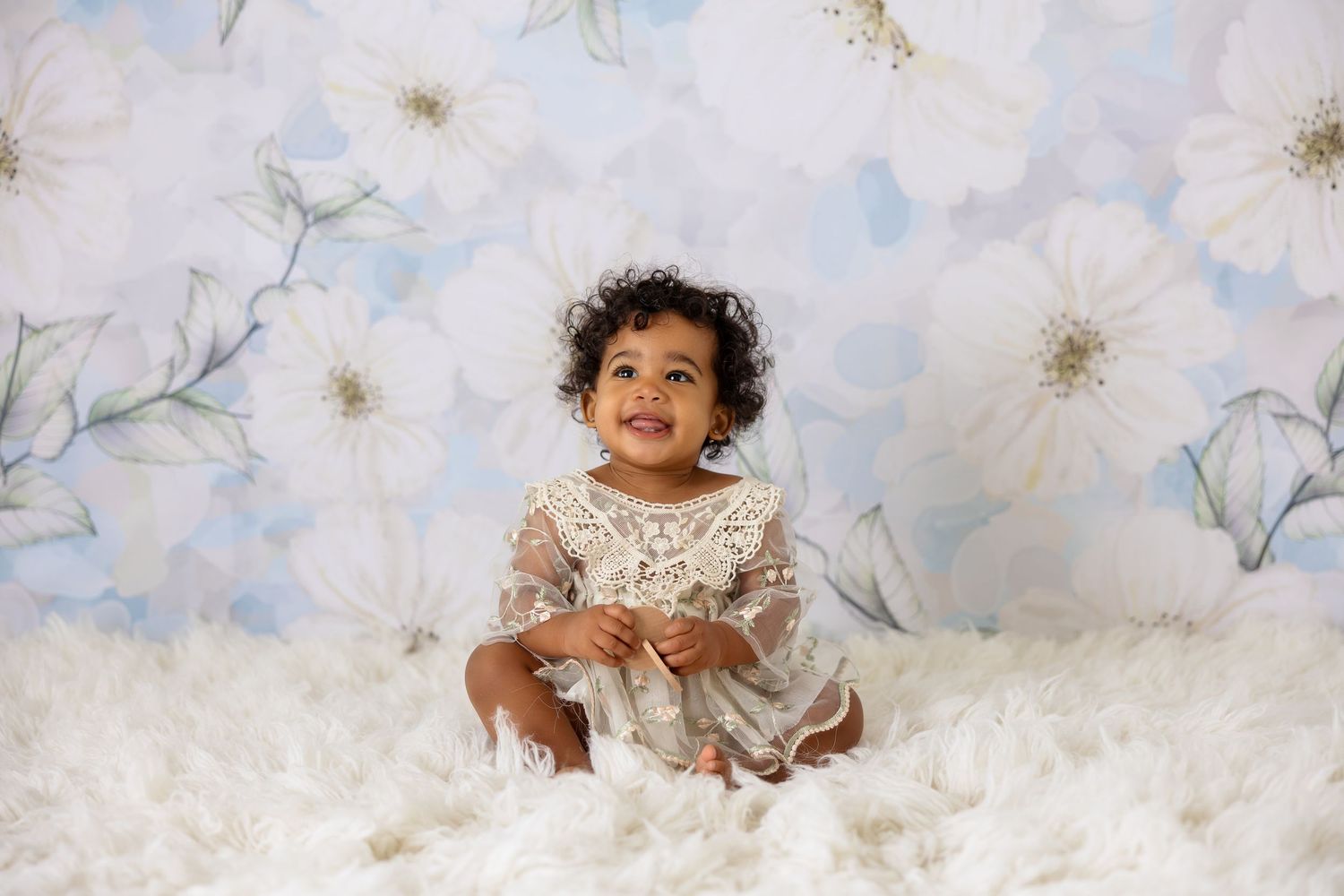 Baby wearing white lace dress poses with cloudy backdrop.
