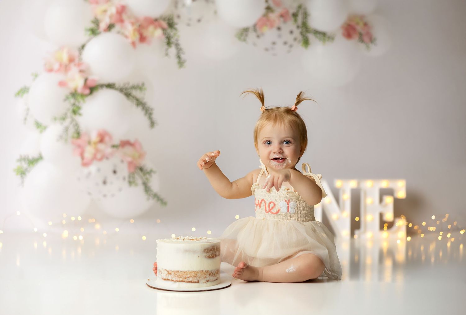 Floral first birthday setup with sparkling lights and illuminated ONE sign against white backdrop.