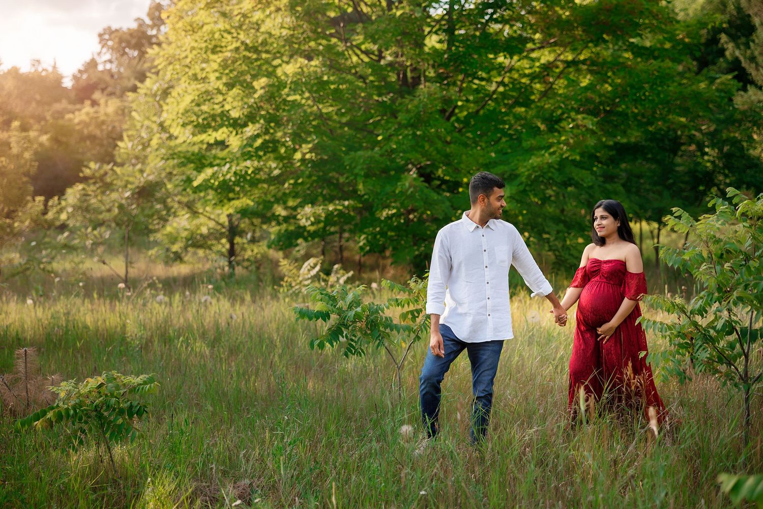 Couple walking together through a sunlit meadow with lush green trees in the background.