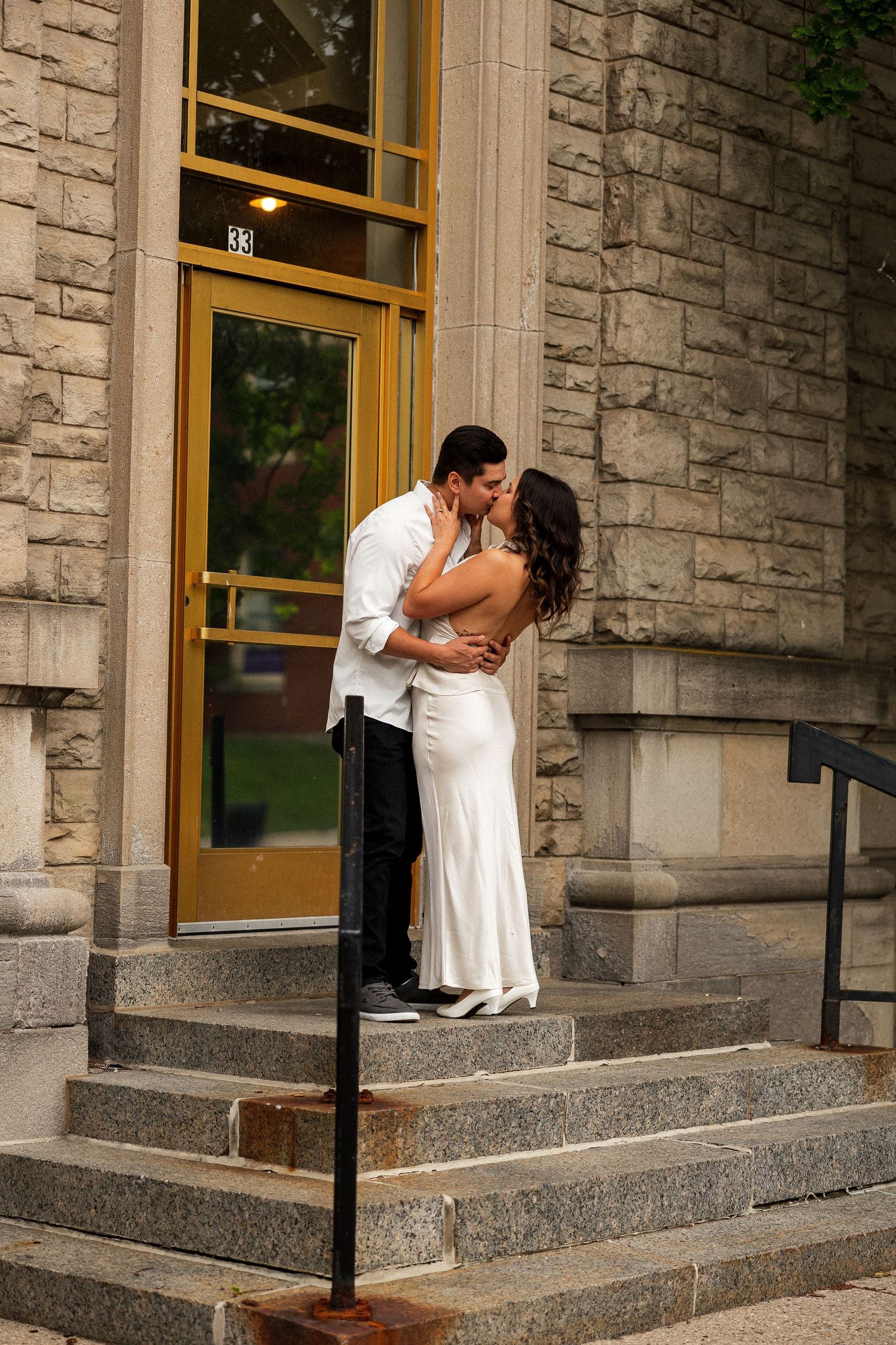 Couple sharing a romantic kiss on stone steps outside a historic building, woman in white dress.
