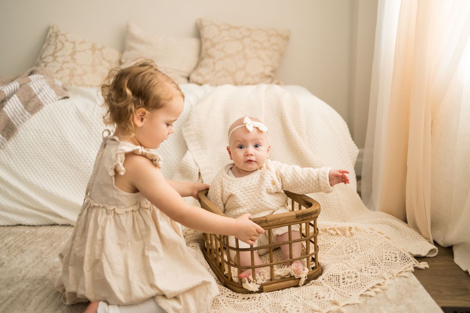 Two children in white dresses play together on a light-colored bed with a woven basket between them.