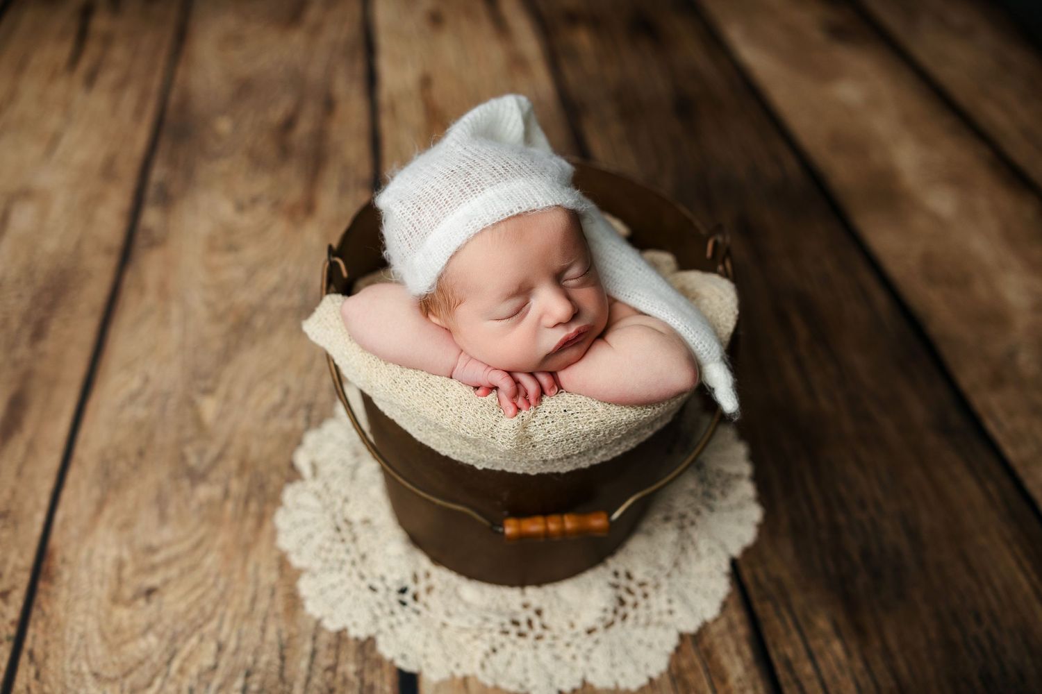 Sleeping newborn wrapped in white blanket rests in brown bowl on lace doily.