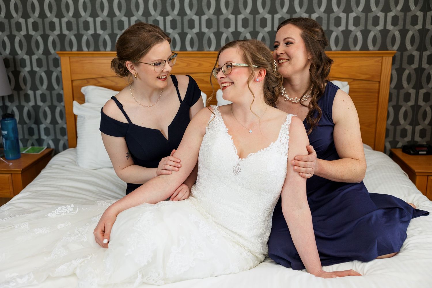 Three women in navy and white dresses share a joyful moment on a bed.
