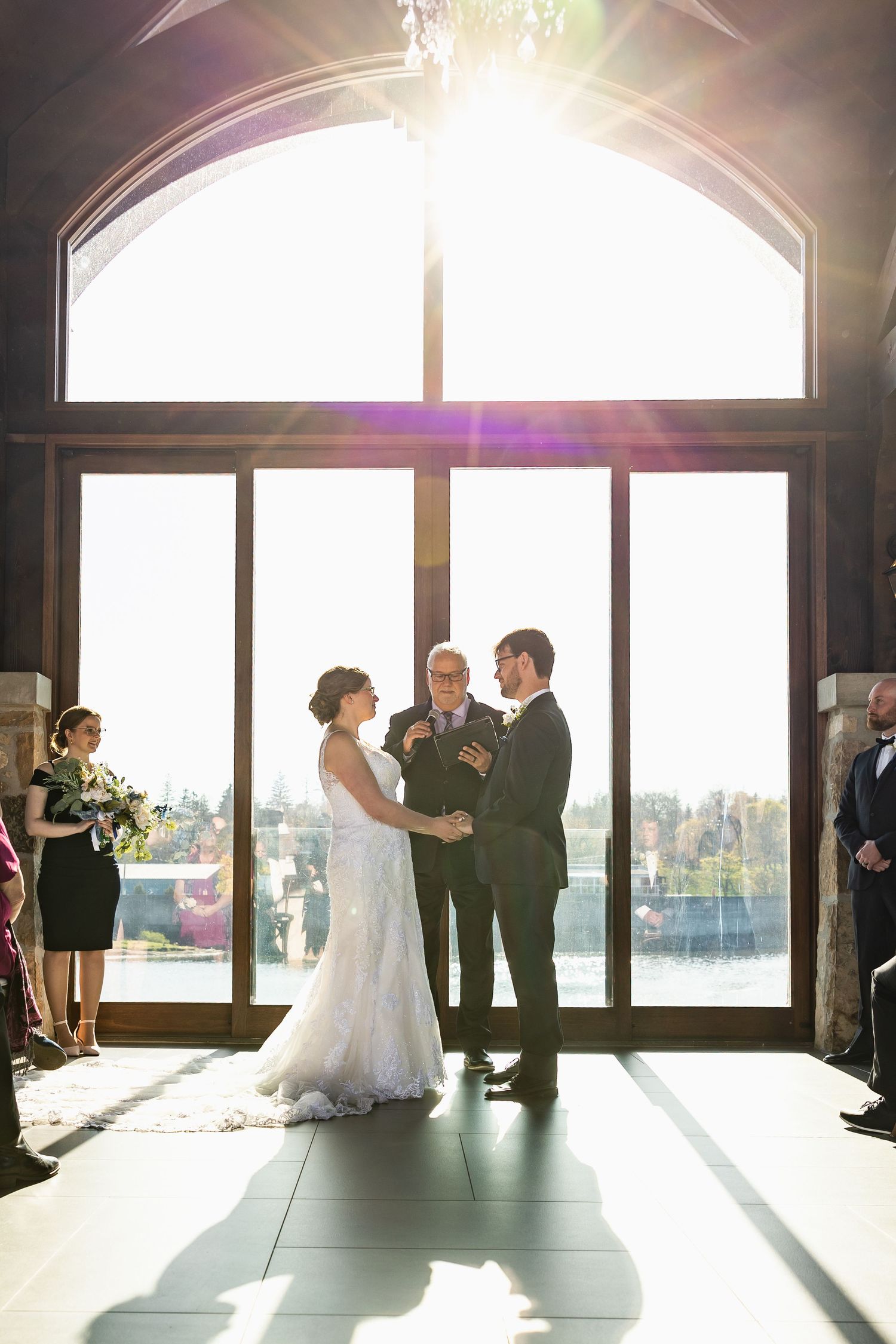 Wedding ceremony silhouetted against bright sunlight streaming through large arched windows.