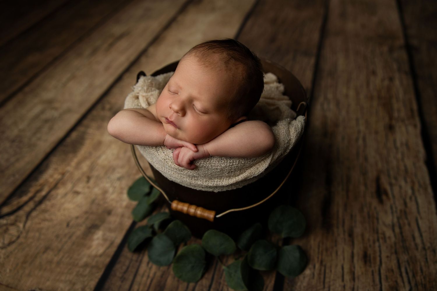 Sleeping baby in bowl surrounded by eucalyptus leaves on wooden surface during studio photoshoot.