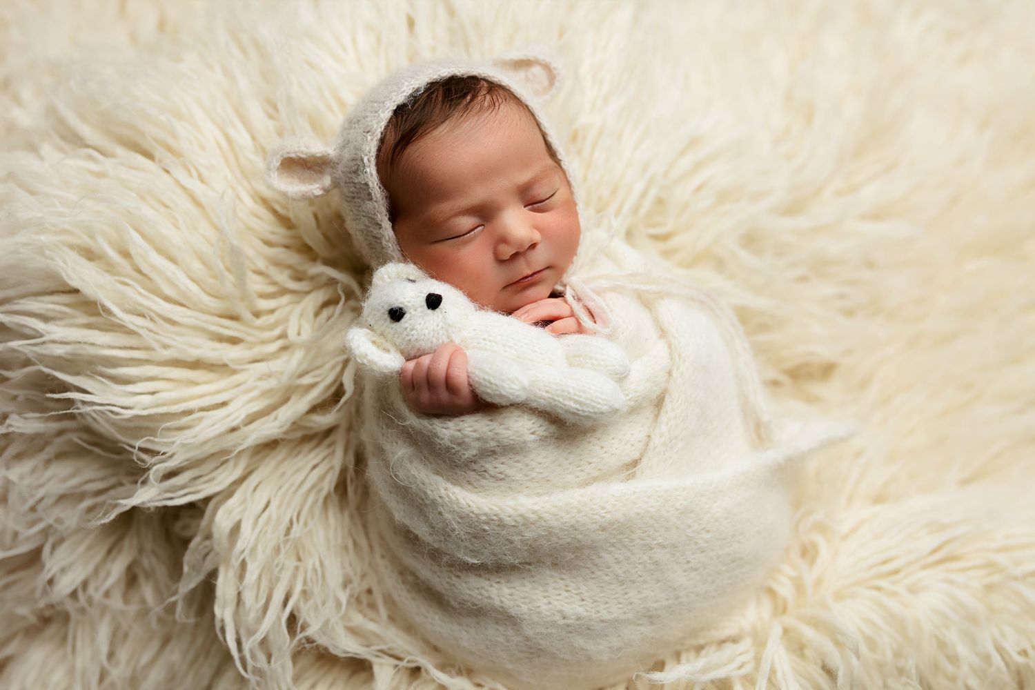 Sleeping newborn wrapped in white swaddle holding small teddy bear on fluffy cream fur backdrop.